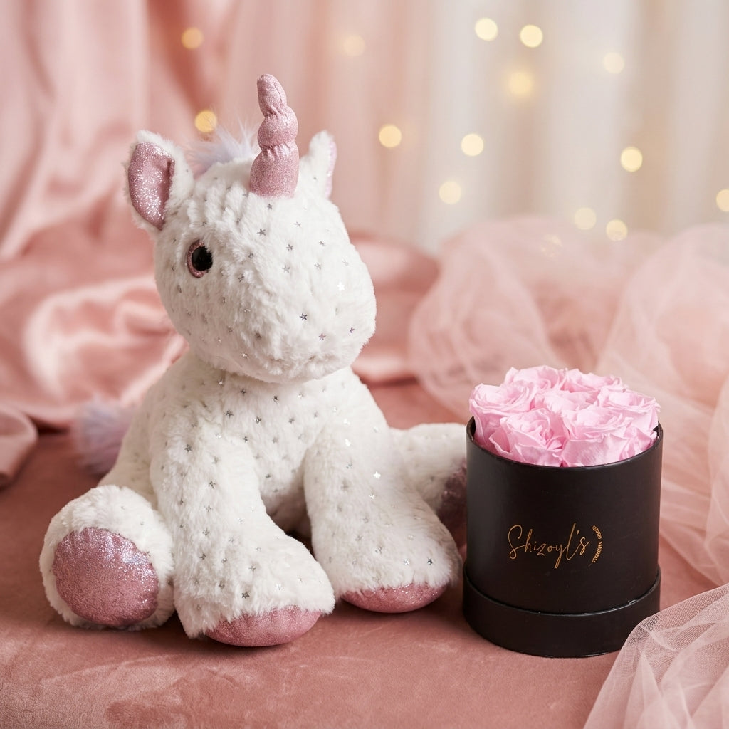 White unicorn plush toy with pink accents next to a black container of pink flowers on a soft pink background.