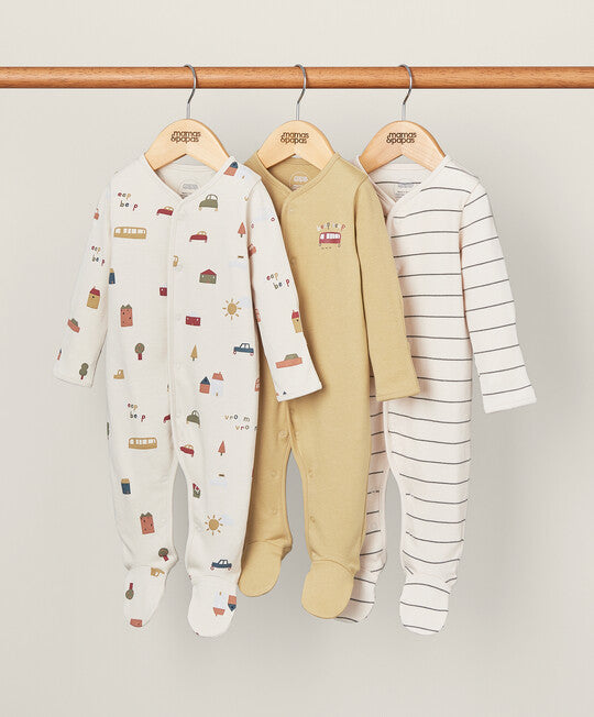 Three baby footed sleepers hang on wooden hangers from a wooden rod against a light beige background. The left sleeper is off-white with a colorful house and sun print.
