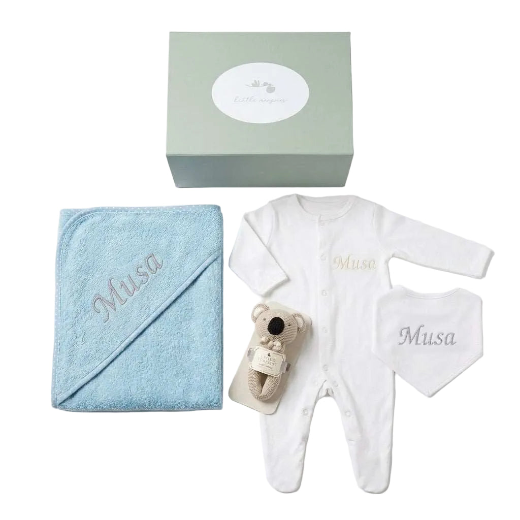 A baby gift set arranged on a white background, including a light blue hooded towel embroidered with 'Musa', a white long-sleeve footed romper, a white bib, a small plush koala toy, and a sage green gift box with a white oval label.