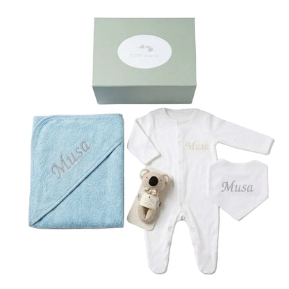 A baby gift set arranged on a white background, including a light blue hooded towel embroidered with 'Musa', a white long-sleeve footed romper, a white bib, a small plush koala toy, and a sage green gift box with a white oval label.